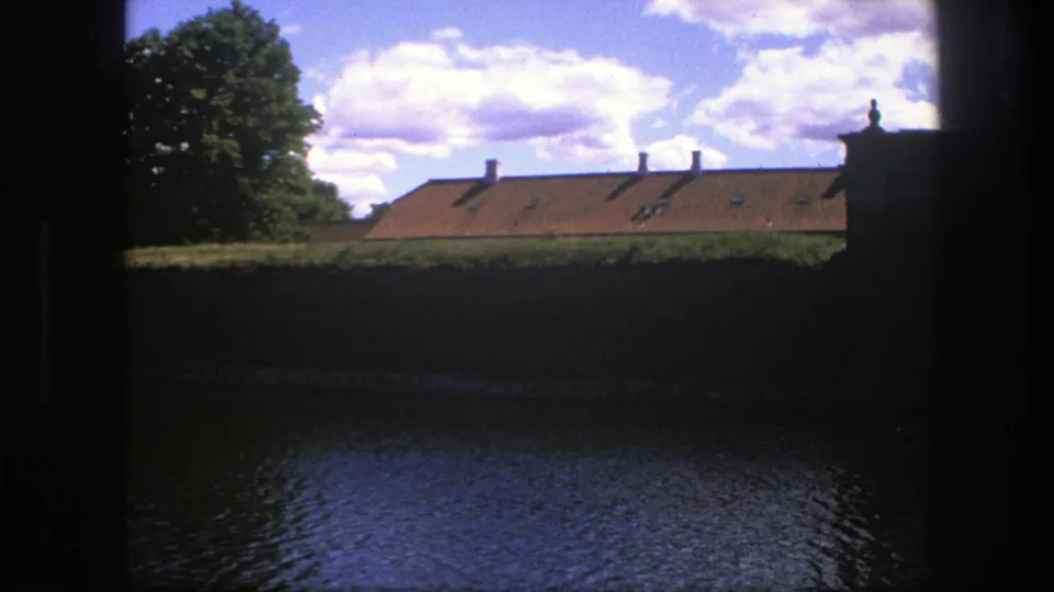 1975 Copenhagen 8mm frame with water in the foreground, a dark wall, long orange roof, trees and bright cloud cover.