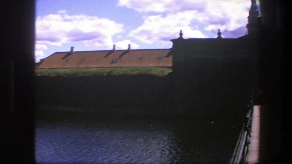 Late-frame 1975 Copenhagen view with rippling water, a dark wall, orange roofline, chimneys and a partial tower or building edge.