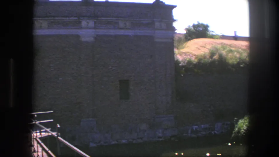The 8mm view pans right from the gateway to a fortress wall beside grass, shrubs, railing, and a raised rampart in Copenhagen.