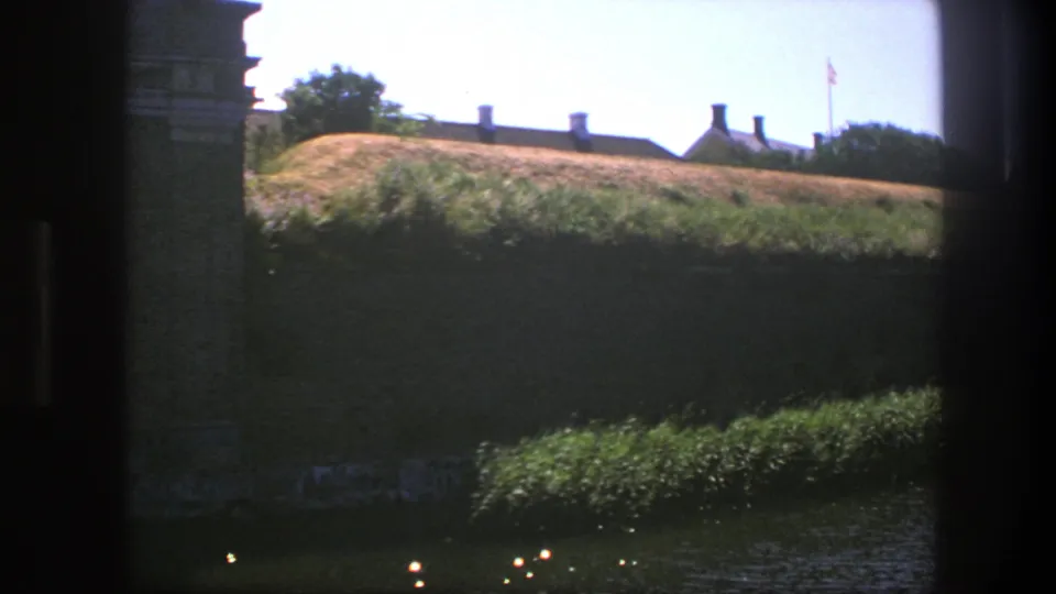 1975 Copenhagen 8mm view across water toward a grassy bank, rooflines, trees, and a distant flag framed by dark edges.