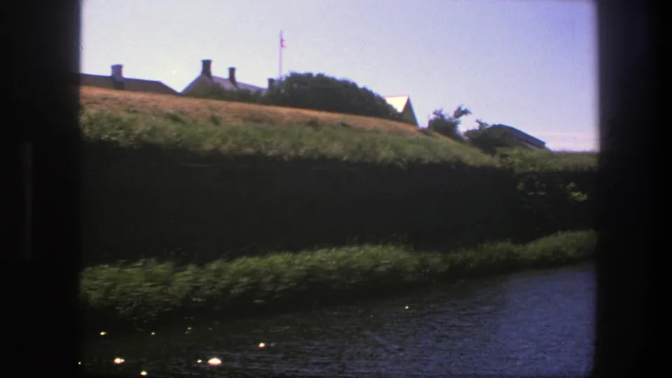 1975 Copenhagen 8mm frame with a dark window or boat edge around water, grassy bank, roof peaks, trees, and open sky.