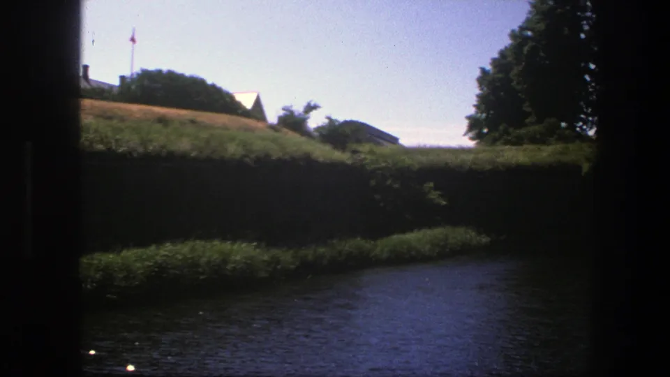 1975 Copenhagen riverside view with rippling water in front, a shadowed bank, grassy slope, trees, roofs, and a small distant flag.