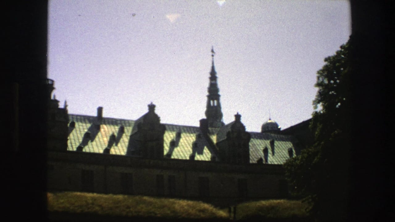 1975 Copenhagen home-movie frame showing a historic building roofline, green roofs, spire, trees, and dark foreground framing.