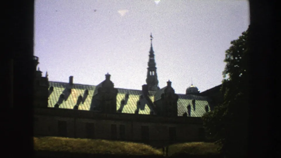 1975 Copenhagen home-movie frame showing a historic building roofline, green roofs, spire, trees, and dark foreground framing.