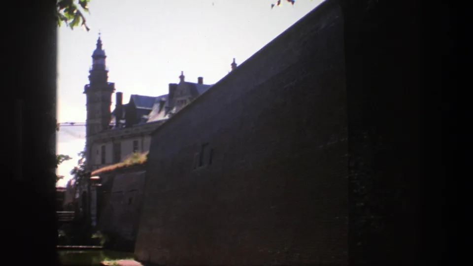 1975 Copenhagen home-movie view along a brick wall toward a historic building with a tower, roofline, trees, and bright sky.