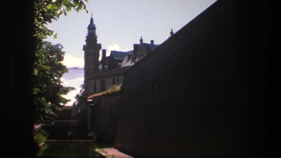 1975 Copenhagen home-movie frame showing a castle tower beyond leafy trees and a shadowed brick wall.