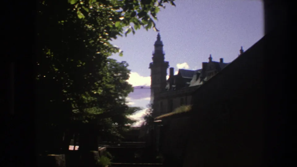 A 1975 8mm frame shows a Copenhagen castle tower and rooftops beyond a dark foreground wall and trees.