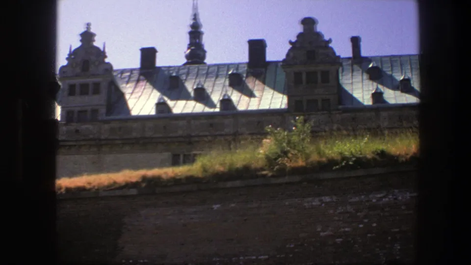 1975 Copenhagen home-movie frame with a green roof, decorative gables, chimneys, grass, and brick wall seen through dark edges.