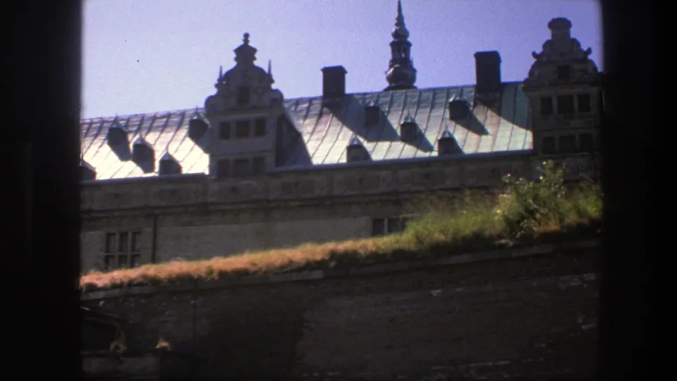 1975 Copenhagen travel footage frame of a sunlit metal roof with decorative gables, chimneys, and a spire beyond dark side framing.
