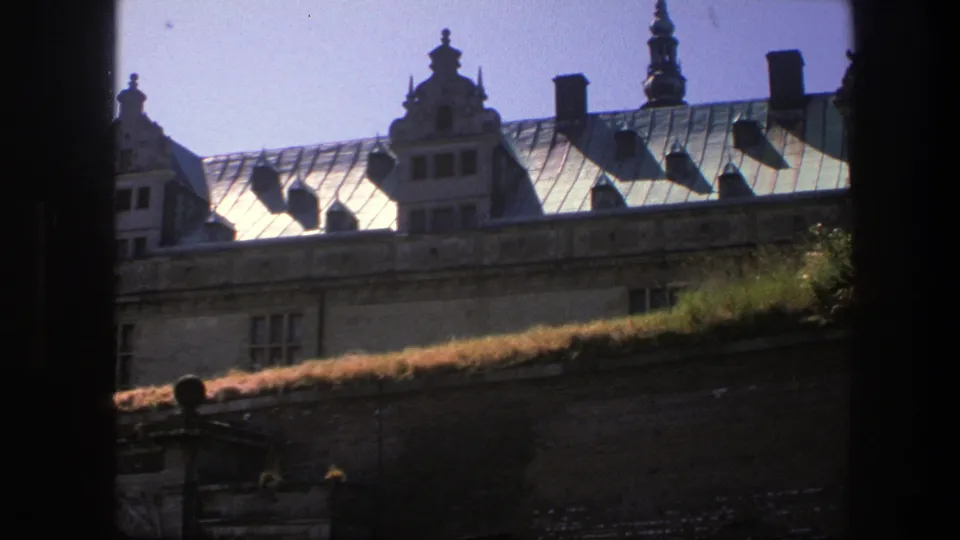 1975 Copenhagen frame showing a central ornate gable, chimneys, metal roof panels, stone facade windows, and a grassy lower ledge.