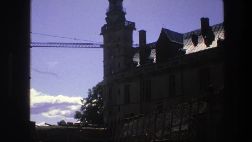 1975 Copenhagen home-movie frame showing the castle tower and roofline in dark backlight, with clouds, trees, and a crane beyond.