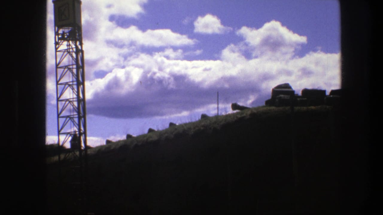Silhouetted metal tower beside a dark ridge under bright clouds and blue sky.