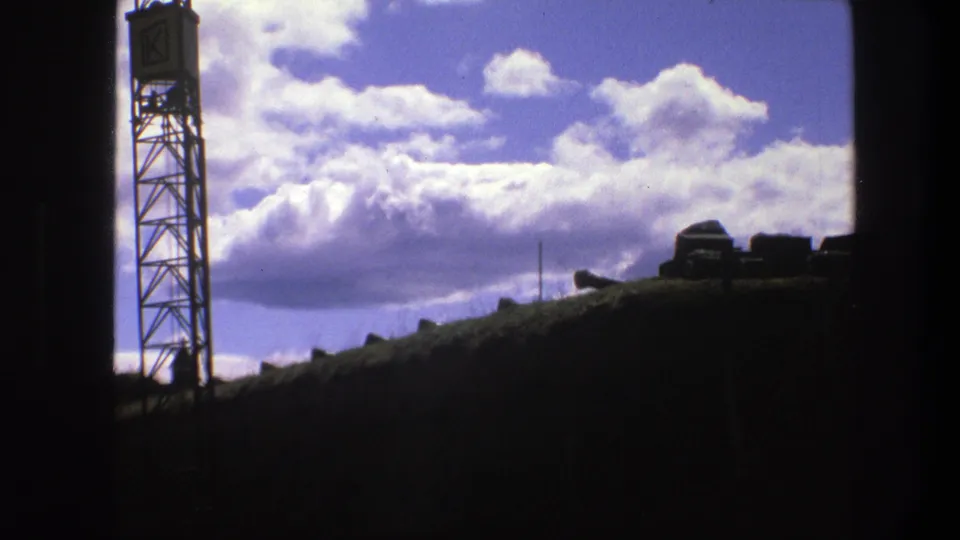 1975 Copenhagen 8mm still of a silhouetted lattice tower, low dark structures, and layered white clouds.