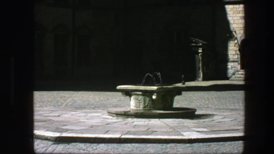 1975 Copenhagen home-movie frame showing a stone fountain flowing in a sunlit courtyard seen from deep shade.