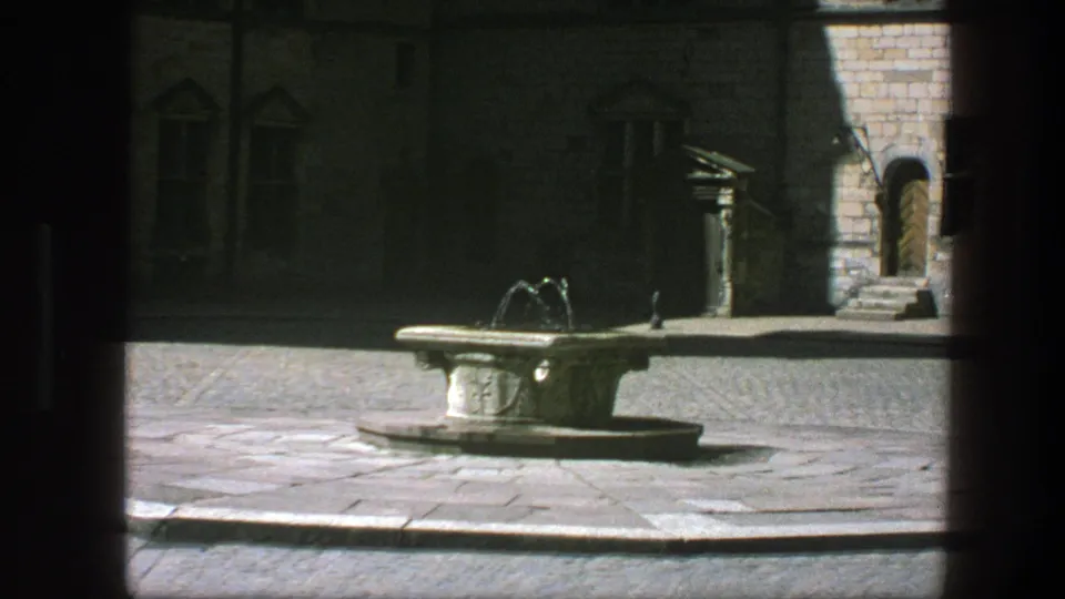 A shaded 8mm frame of the courtyard fountain, with water arcs, paving, and an arched doorway in the background.