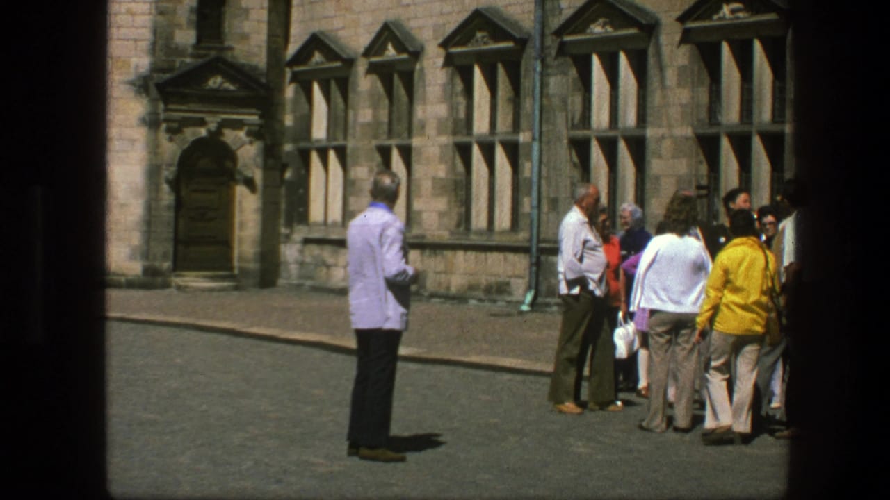 Adults gather outside a stone building while a man in a light purple shirt stands apart smoking.