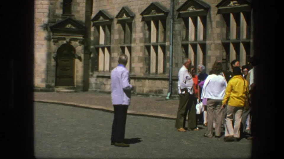 1975 Copenhagen home-movie frame showing adults near a historic stone facade, with one man standing separately in bright daylight.