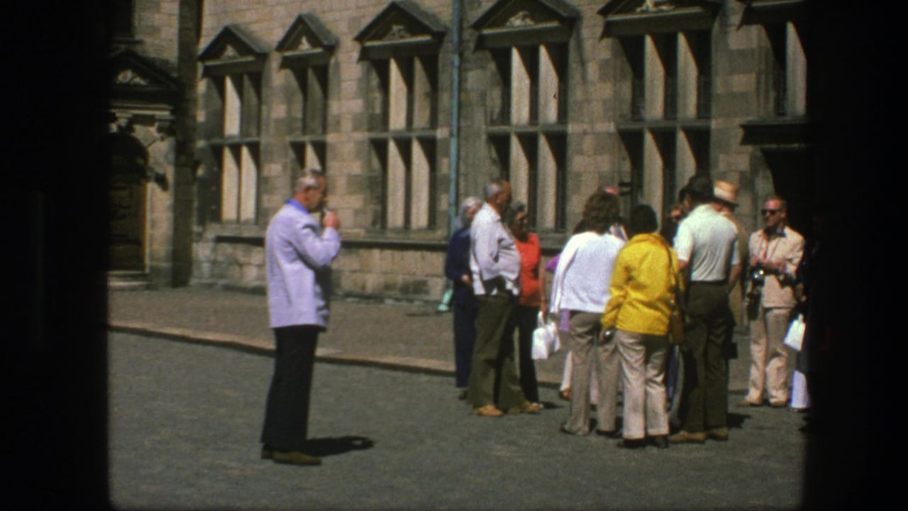 A cluster of adults talks beside tall windows as a man in a light purple shirt smokes nearby.