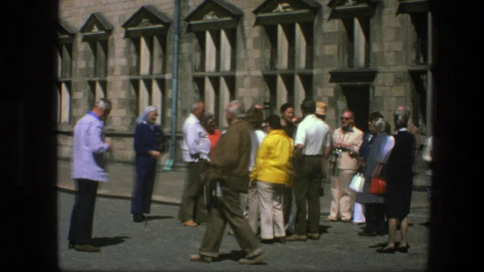 1975 Copenhagen frame with adults gathered outside a stone building and a man crossing through the foreground.