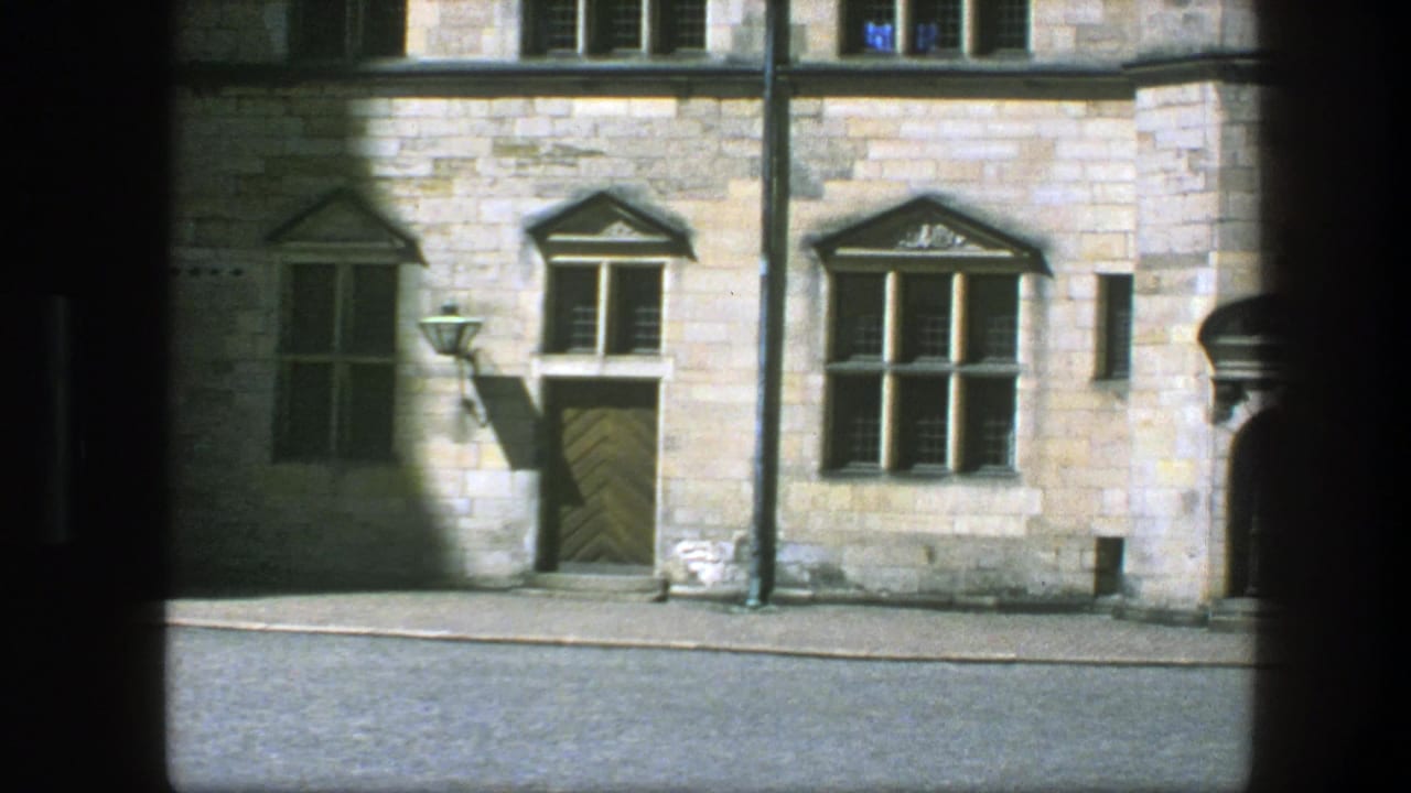Stone facade with paired windows, wooden door, street lamp, and sunlit wall sections.