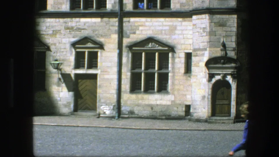 1975 Copenhagen 8mm frame with a stone facade, arched doorway, mullioned windows, street lamp, and a partially visible passerby at the lower edge.
