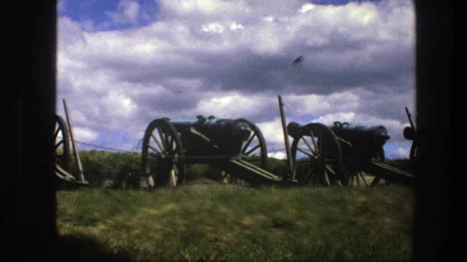 1975 Copenhagen 8mm frame of historic cannon carriages lined on grass, with dark foreground framing and a clouded sky overhead.