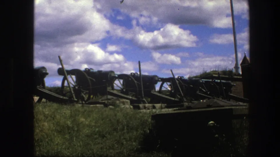 1975 Copenhagen home-movie frame with historic artillery on grass, a tall pole in the background, and dark foreground framing.