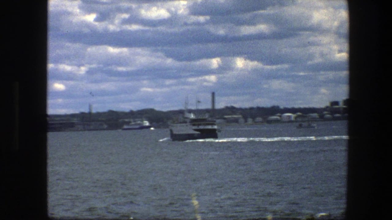 Small ferry moving away across harbor water under a cloudy sky.