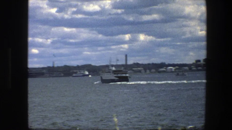 1975 Copenhagen harbor view of a small ferry departing, framed by dark window edges with distant boats and industrial shoreline.
