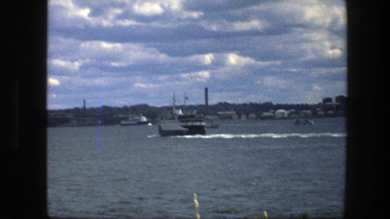 Small ferry continues away through Copenhagen harbor in 1975, with smokestacks and low waterfront buildings on the horizon.