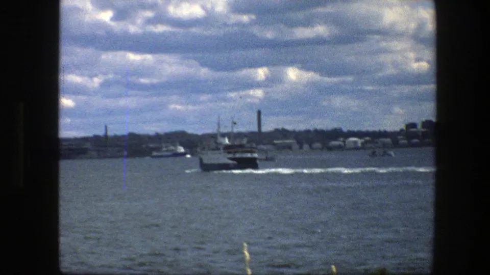Small ferry continues away through Copenhagen harbor in 1975, with smokestacks and low waterfront buildings on the horizon.