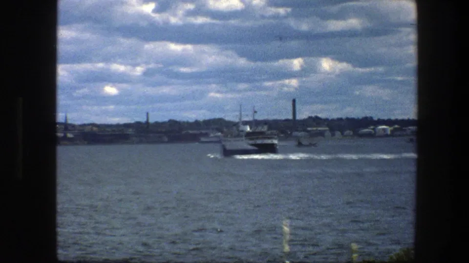 1975 Copenhagen harbor footage shows a small ferry departing across open water, framed by dark edges and a cloudy industrial skyline.