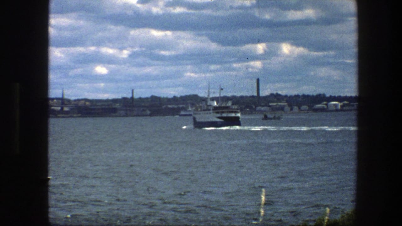 1975 Copenhagen harbor view with a larger ferry on the water, distant shoreline structures, cloud cover, and dark frame edges from the filming position.
