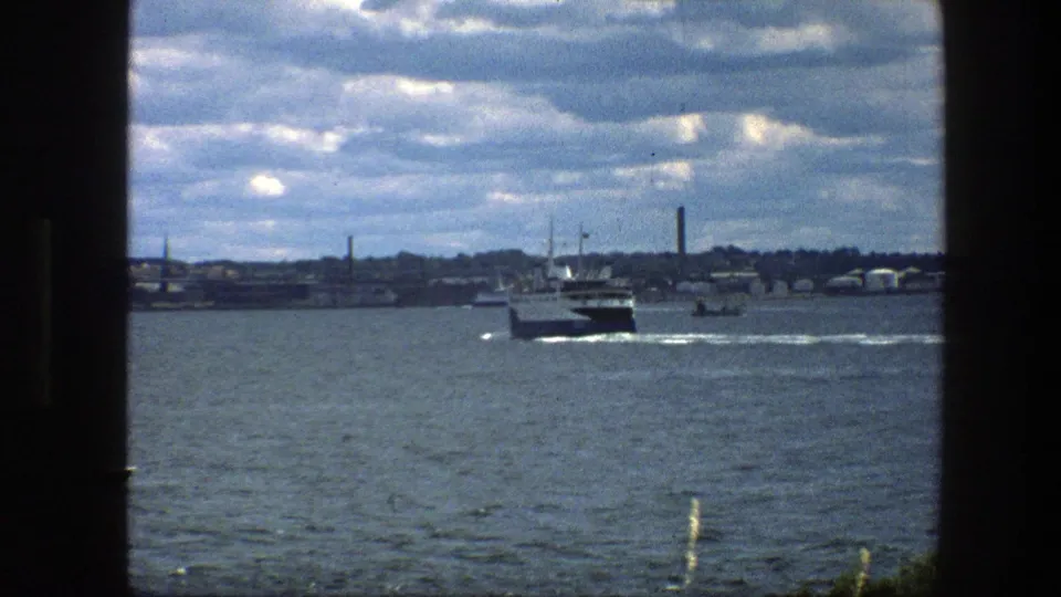 1975 Copenhagen harbor view with a larger ferry on the water, distant shoreline structures, cloud cover, and dark frame edges from the filming position.