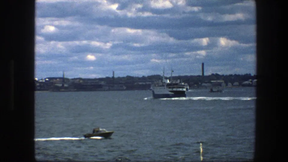 Copenhagen harbor scene with a small motorboat in the foreground, a larger ferry beyond it, white wakes, and a low shoreline under clouds.