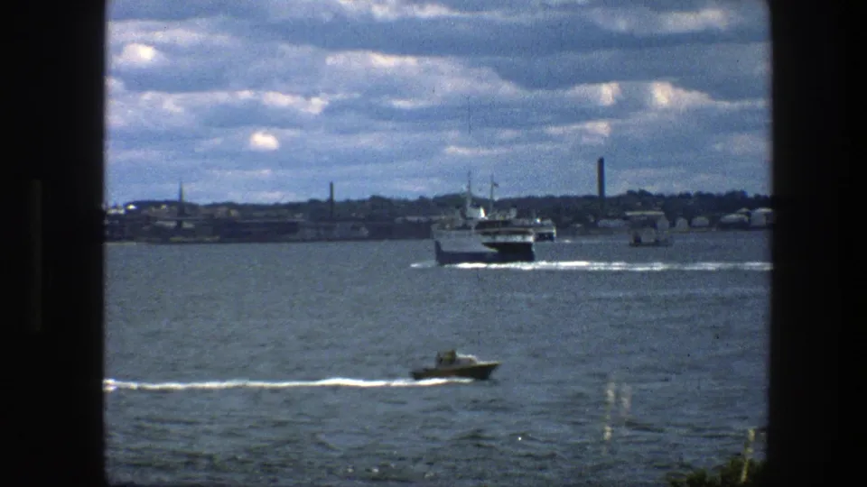 1975 Copenhagen waterfront footage showing a small foreground boat, a larger ferry mid-distance, low harbor structures, and textured 8mm color.