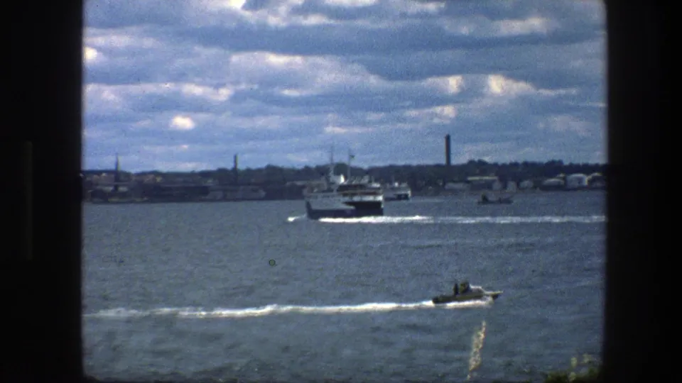 Copenhagen harbor view with a ferry mid-frame, a small motorboat at lower right, distant waterfront structures, and dark 8mm frame edges.