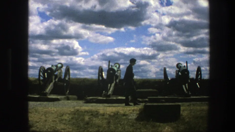 A dark-clothed man walks across a grassy cannon display in Copenhagen, with heavy clouds filling the background sky.