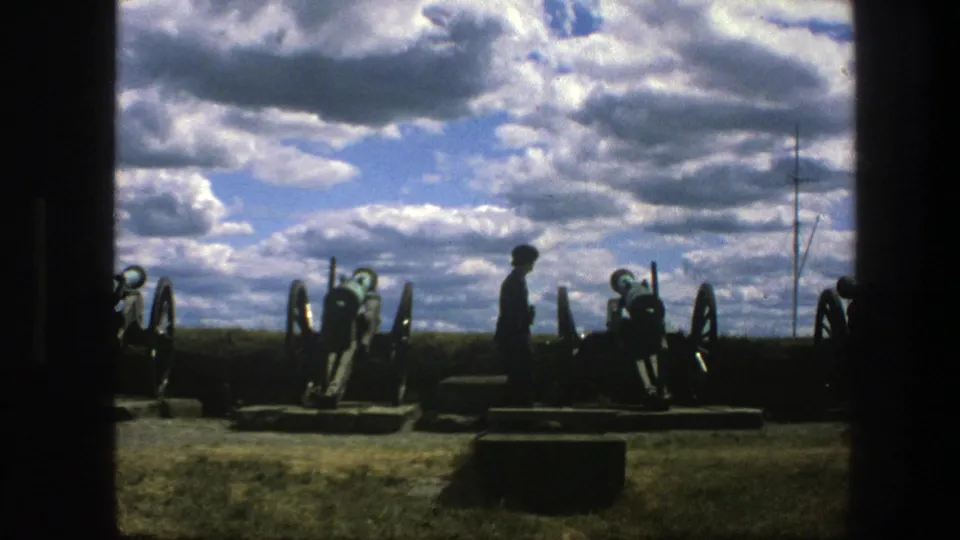 The visitor is silhouetted near historic cannons at the Copenhagen site, with a bright cloud bank and dark film border visible.