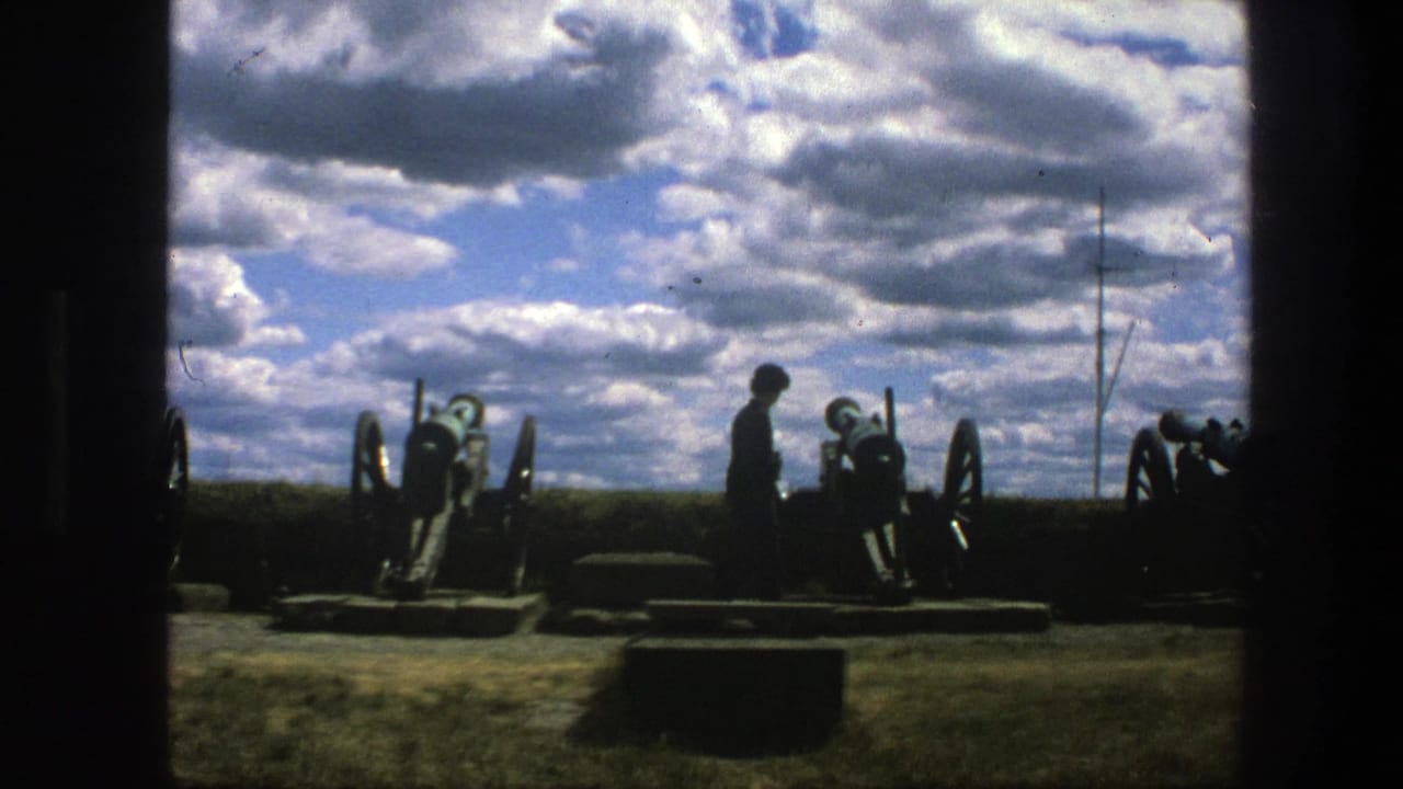 Silhouetted person stands among historic cannons beneath a cloud-filled sky.