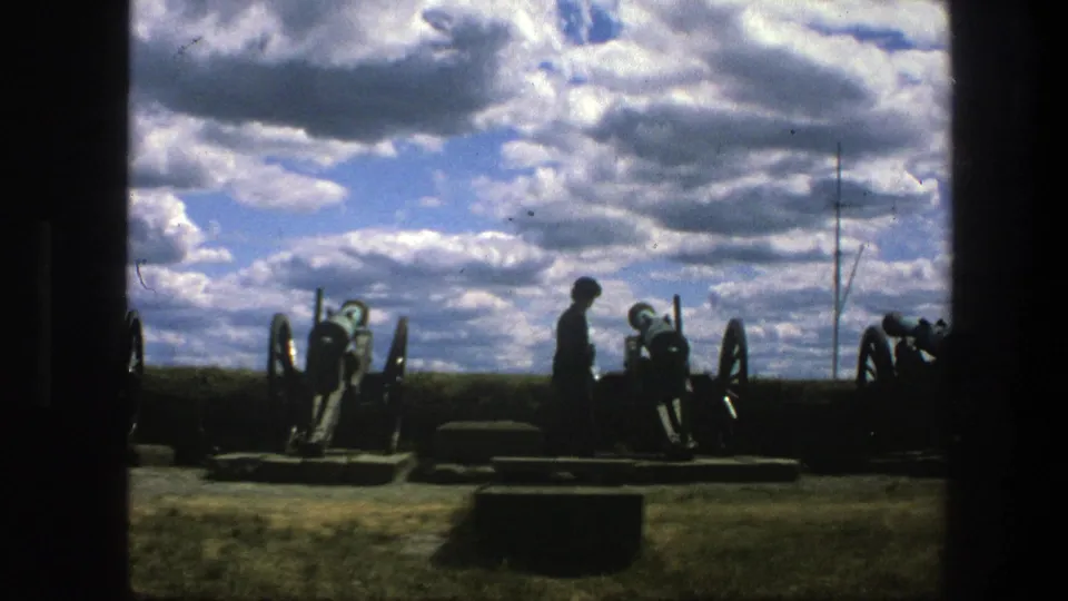 1975 8mm Copenhagen home-movie frame showing a silhouetted visitor near wheeled cannons, framed by dark doorway edges and open sky.