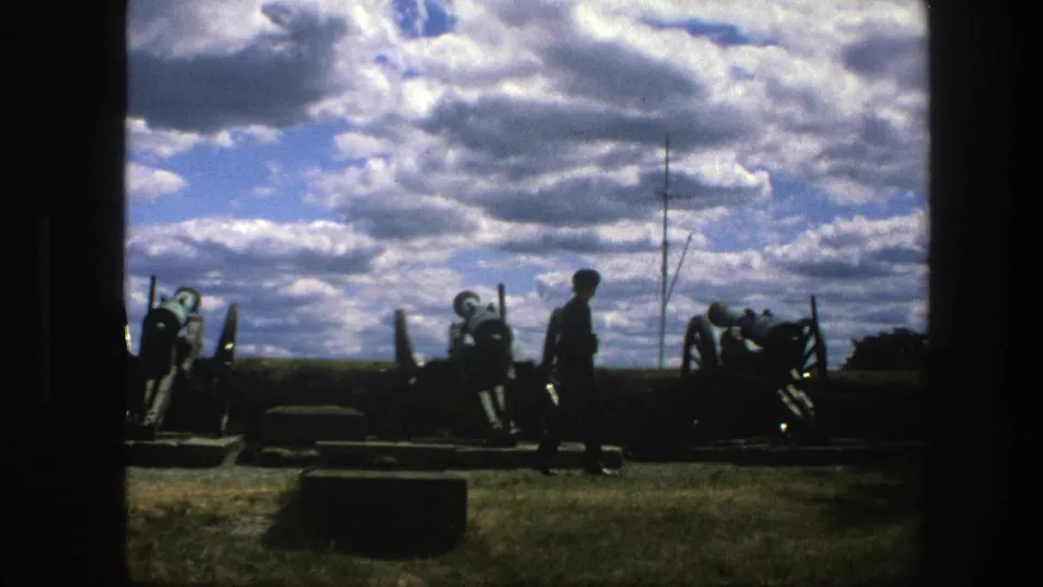 1975 8mm Copenhagen home-movie still showing a visitor in silhouette crossing in front of preserved cannons and a low platform.