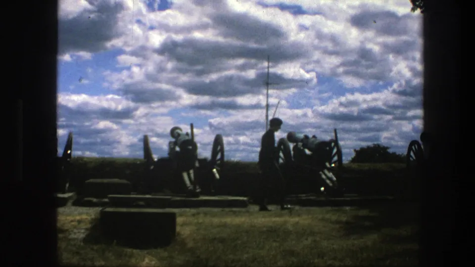 1975 8mm Copenhagen frame of a silhouetted visitor beside historic wheeled cannons, with grass, shadowed edges, and a bright cloud bank.