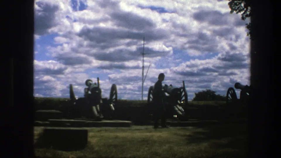 1975 Copenhagen-area 8mm frame with silhouetted cannons, a standing visitor, open water, and dramatic clouds.