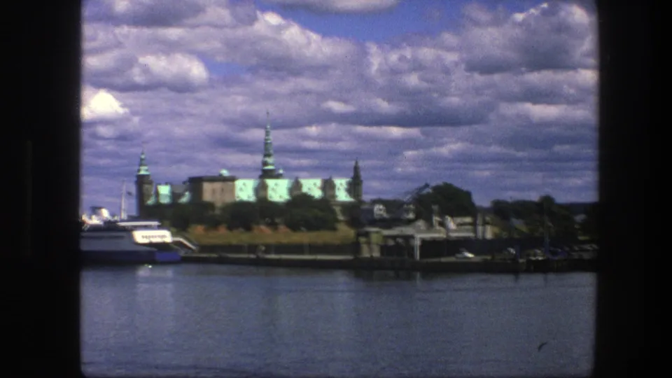 1975 Copenhagen 8mm harbor view with calm water, a ferry, green-roofed buildings, and a broad cloudy sky.
