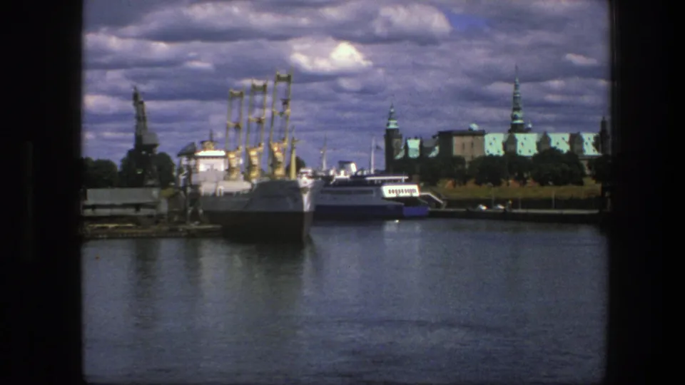 1975 Copenhagen waterfront scene with calm harbor water, docked boats, yellow cranes, and historic rooflines under a cloudy sky.
