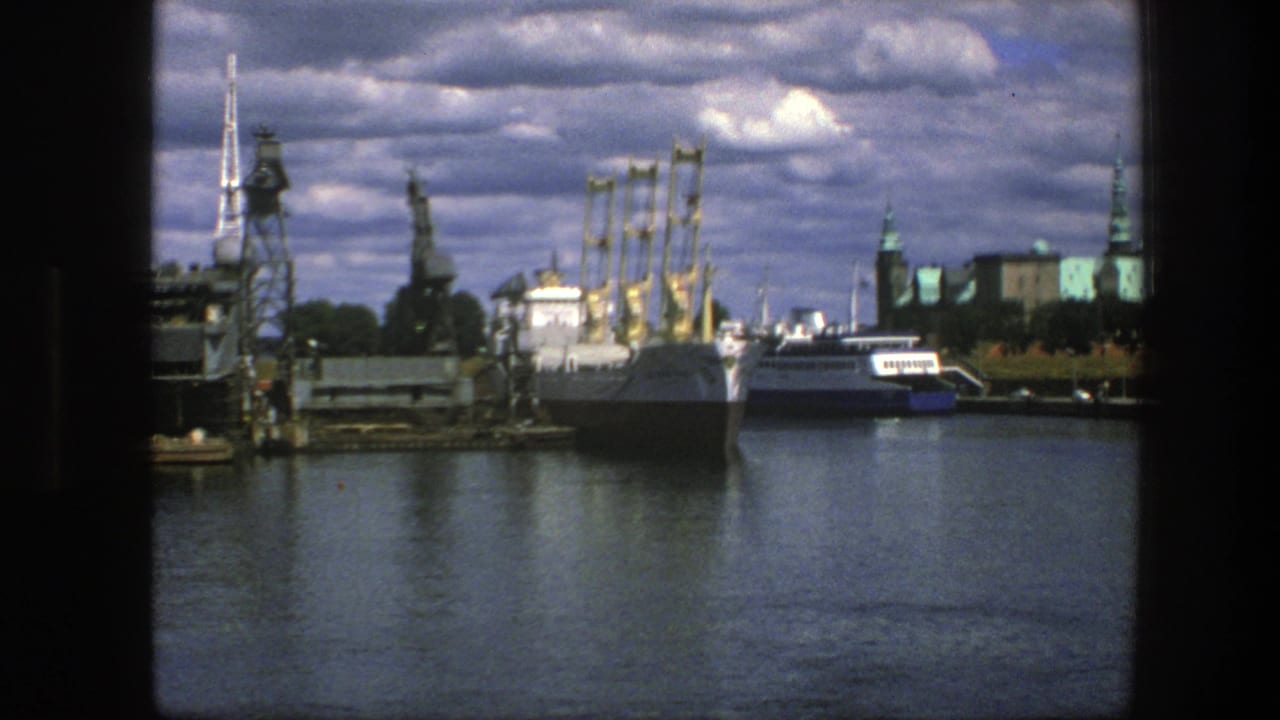 Harbor water with docked vessels, cranes, industrial platforms, and city spires under cloudy sky.