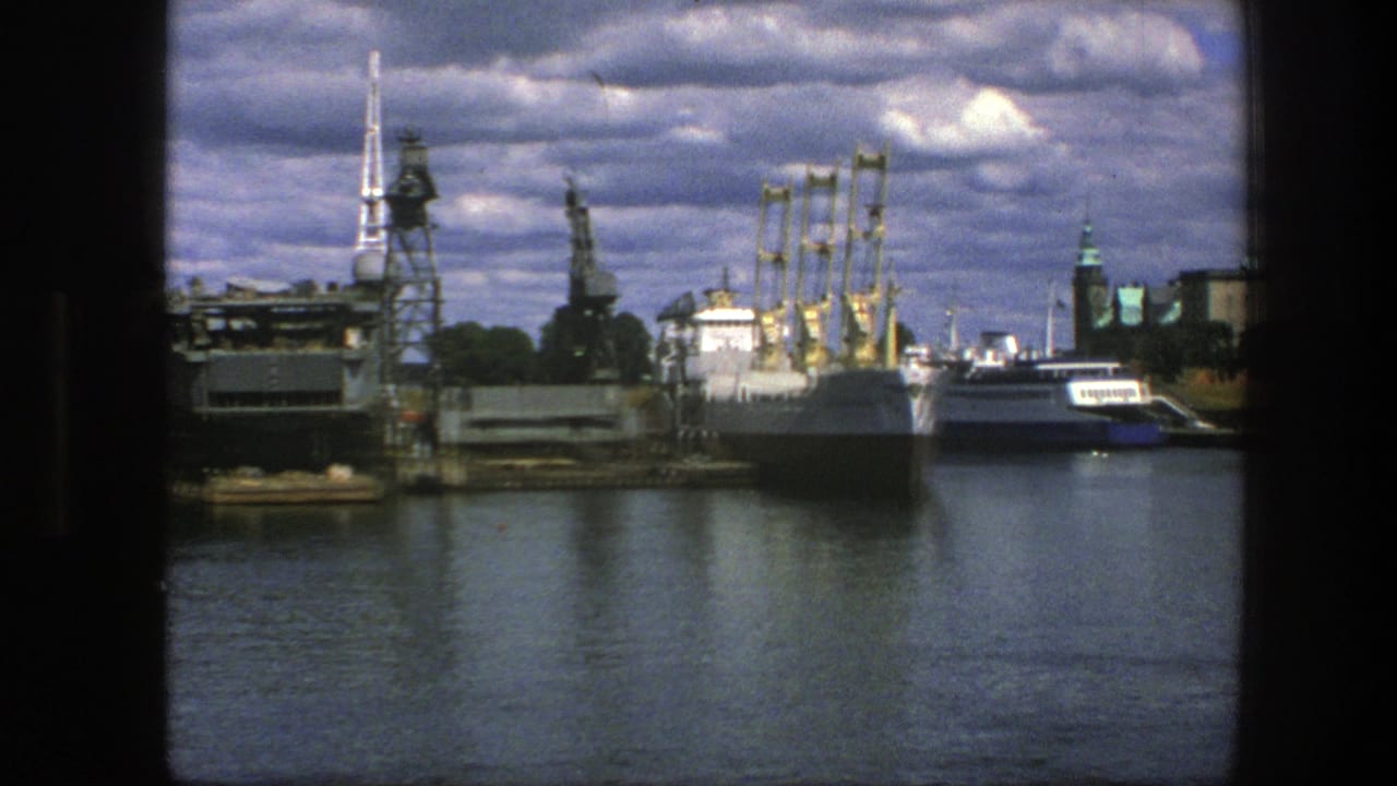 Docked boats and harbor cranes seen across water with spires and heavy clouds behind them.