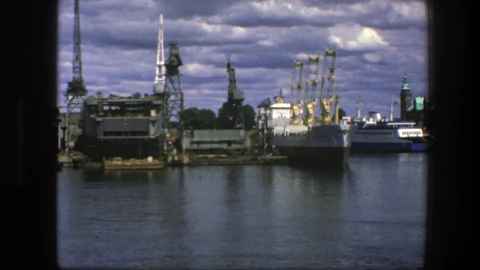 1975 Copenhagen waterfront view with industrial platforms, a docked ship, cranes, harbor water, and city spires in the distance.