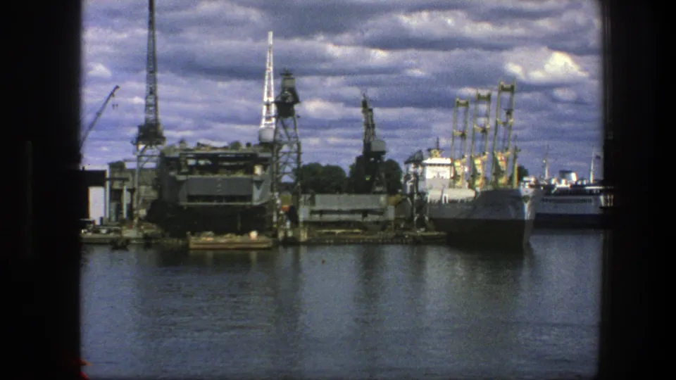 Copenhagen harbor scene from 1975 with cranes, platforms, docked vessels, calm water, and historic spires visible beyond the port.